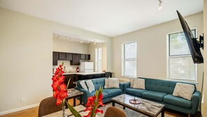 A living room with a sofa, coffee table, and a dining table with chairs. There is a kitchen area in the background.