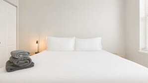 A neatly made bed with white sheets and pillows, a stack of folded towels, and a bedside lamp.