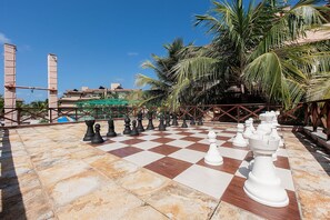 Un tablero de ajedrez grande al aire libre, dispuesto en un patio con baldosas, con palmeras y edificios al fondo.