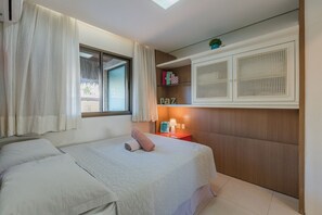 Un dormitorio con cama, mesita de noche, estante con libros y ventana con cortinas.
