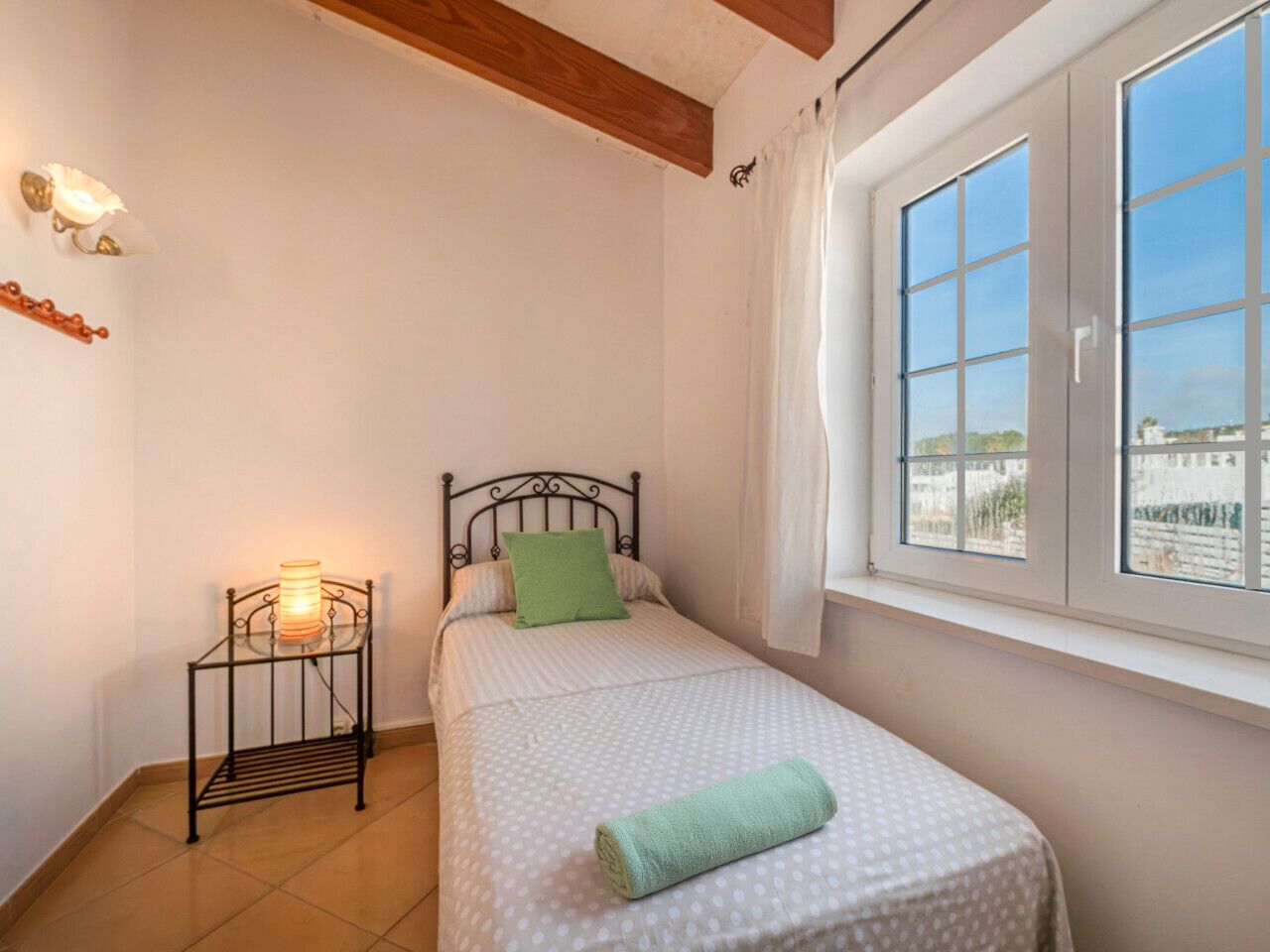 Ein kleines Schlafzimmer mit einem Einzelbett, einem Nachttisch mit Lampe, einem Fenster mit Vorhängen und Blick auf Gebäude.