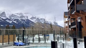 Zona de piscina con vistas a montañas nevadas, una terraza de madera y un edificio moderno con balcones.