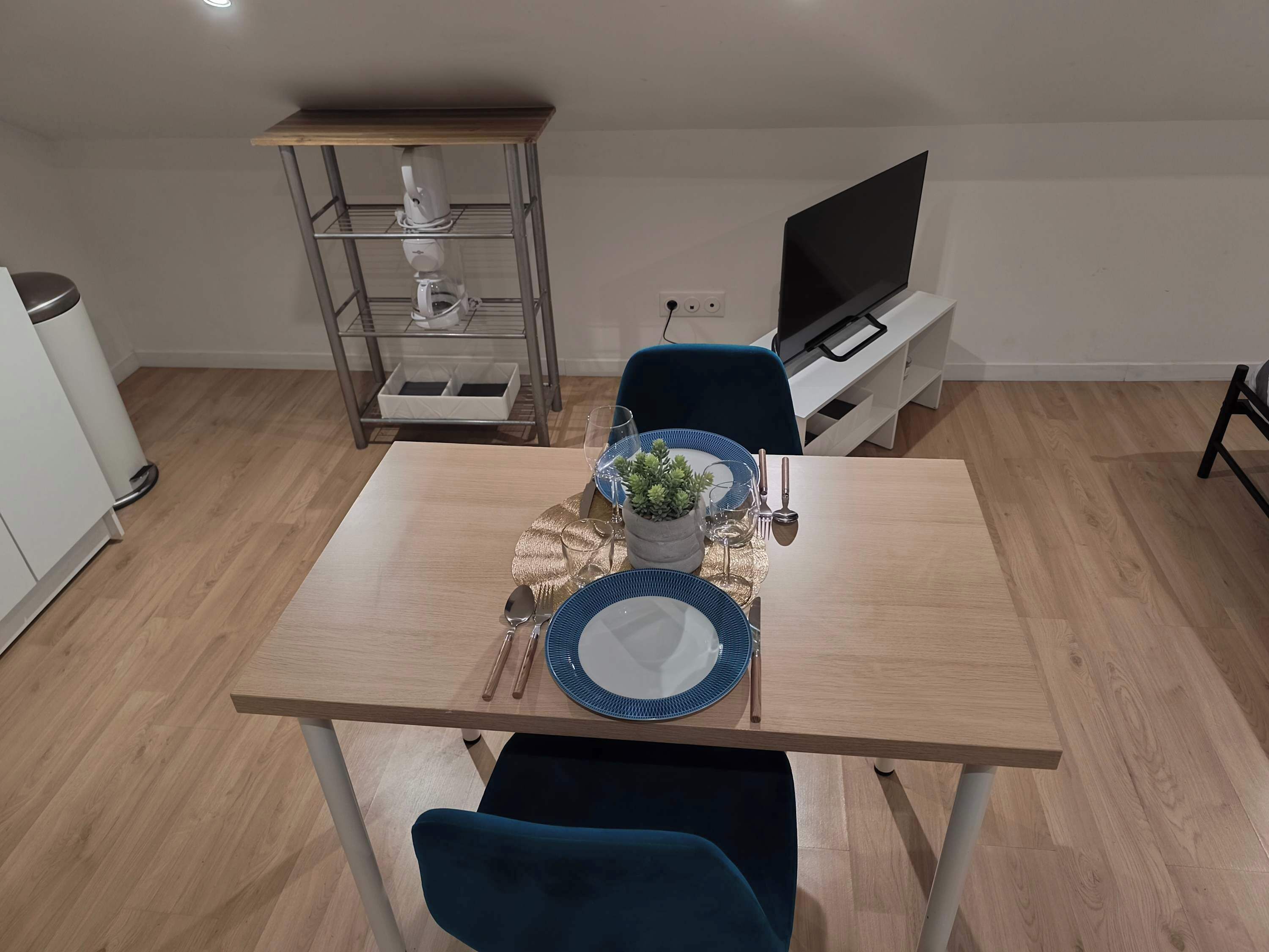 A dining area with a wooden table set for two, a blue chair, a small potted plant, and a TV in the background.
