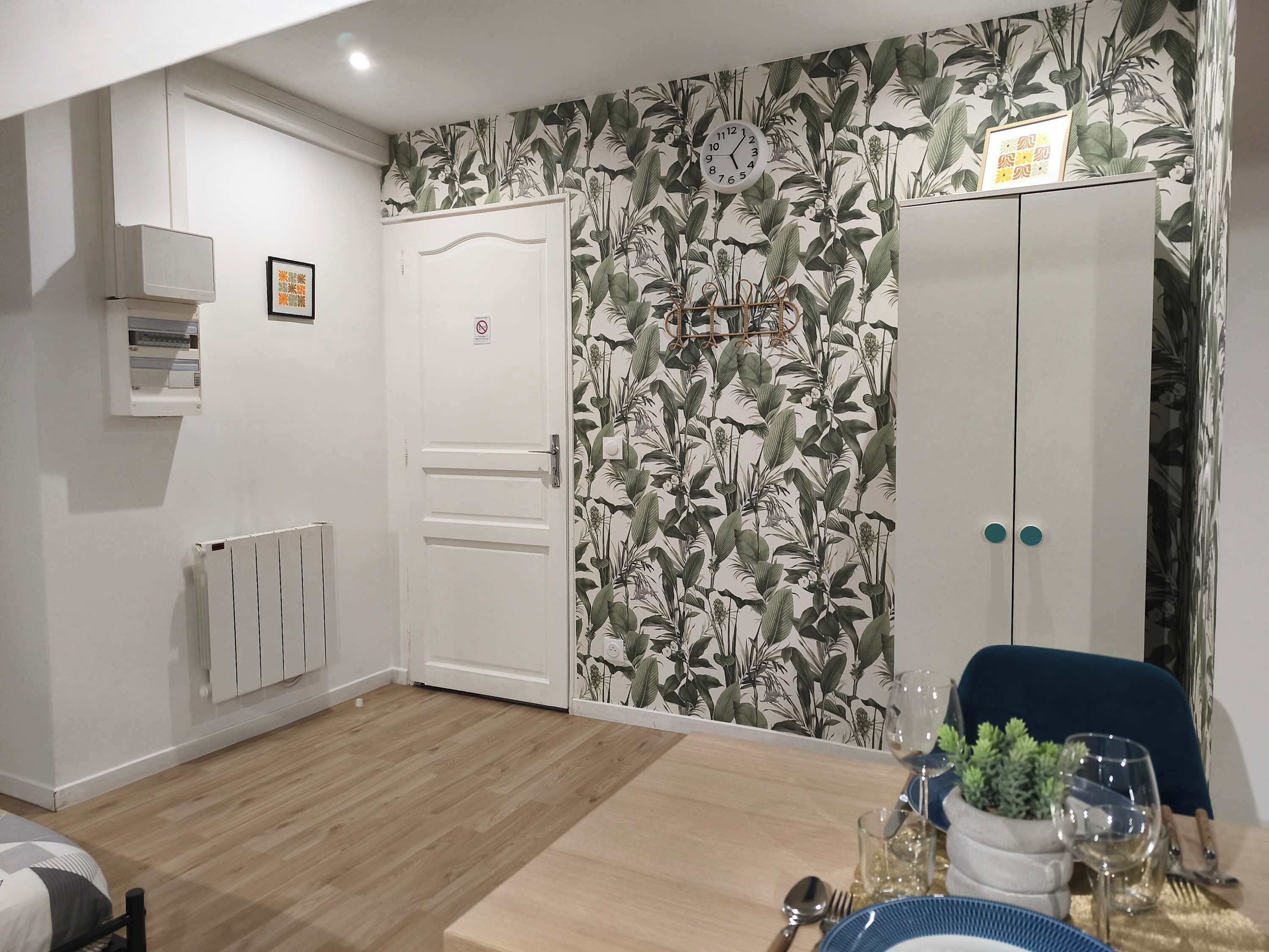 Una habitación con una puerta blanca, un radiador, un reloj en la pared y una mesa de comedor con platos, vasos y una planta.