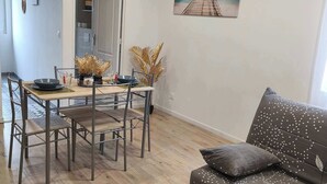 Un salon moderne avec une table à manger en bois, des chaises en métal, un canapé orné d’un motif végétal et un tableau représentant une scène de plage accroché au mur.