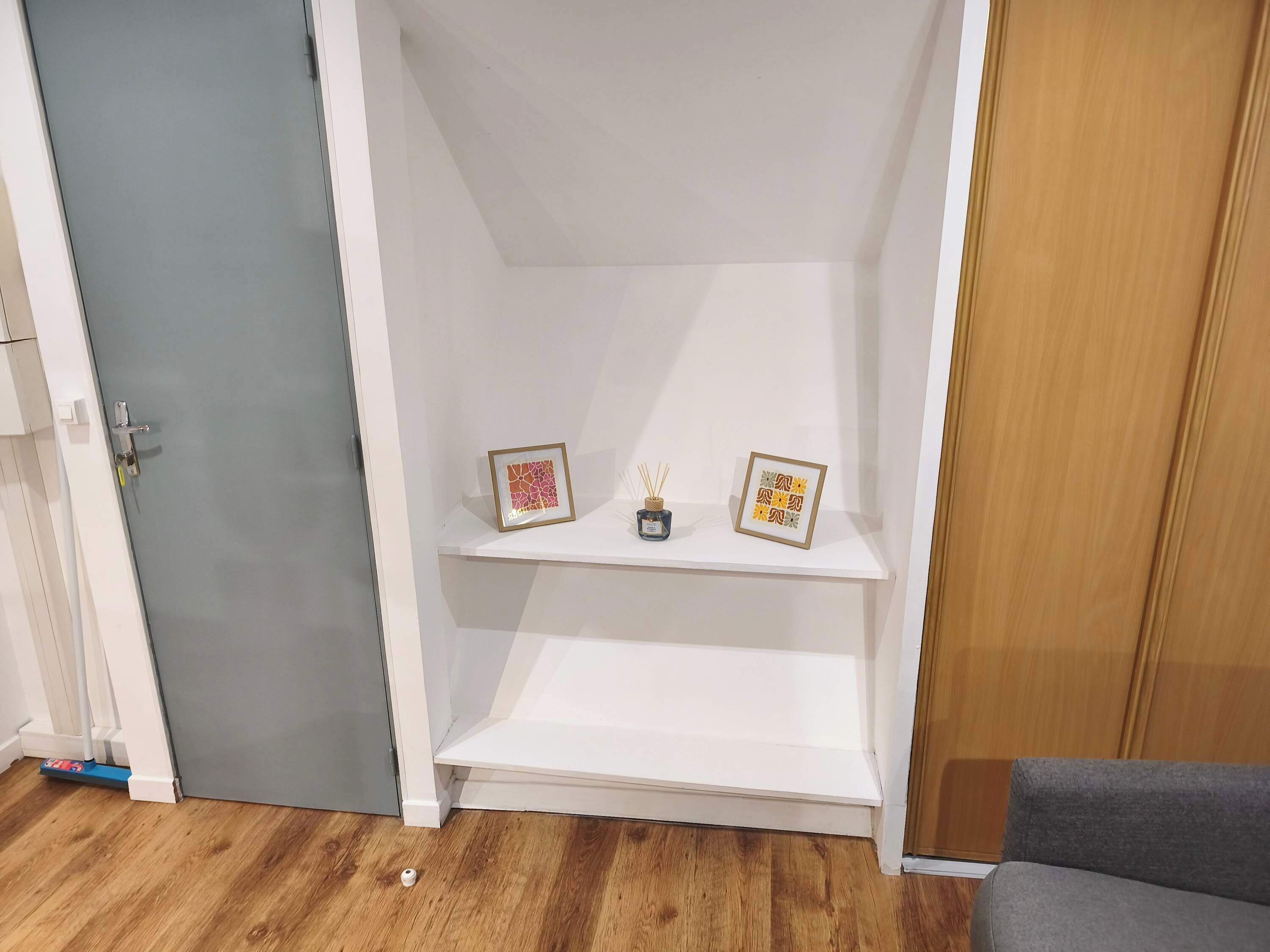Una habitación con una puerta gris, un estante blanco con dos cuadros enmarcados, un pequeño florero con una vela y un sofá gris.