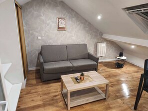 Une chambre de style loft moderne, avec un plafond en pente, un canapé gris, une table basse en bois et une petite table ronde avec une lampe.