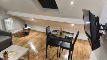 Un espacio habitable compacto con comedor, sofá, mesa de centro, televisor montado en la pared y tragaluz sobre la mesa de comedor.