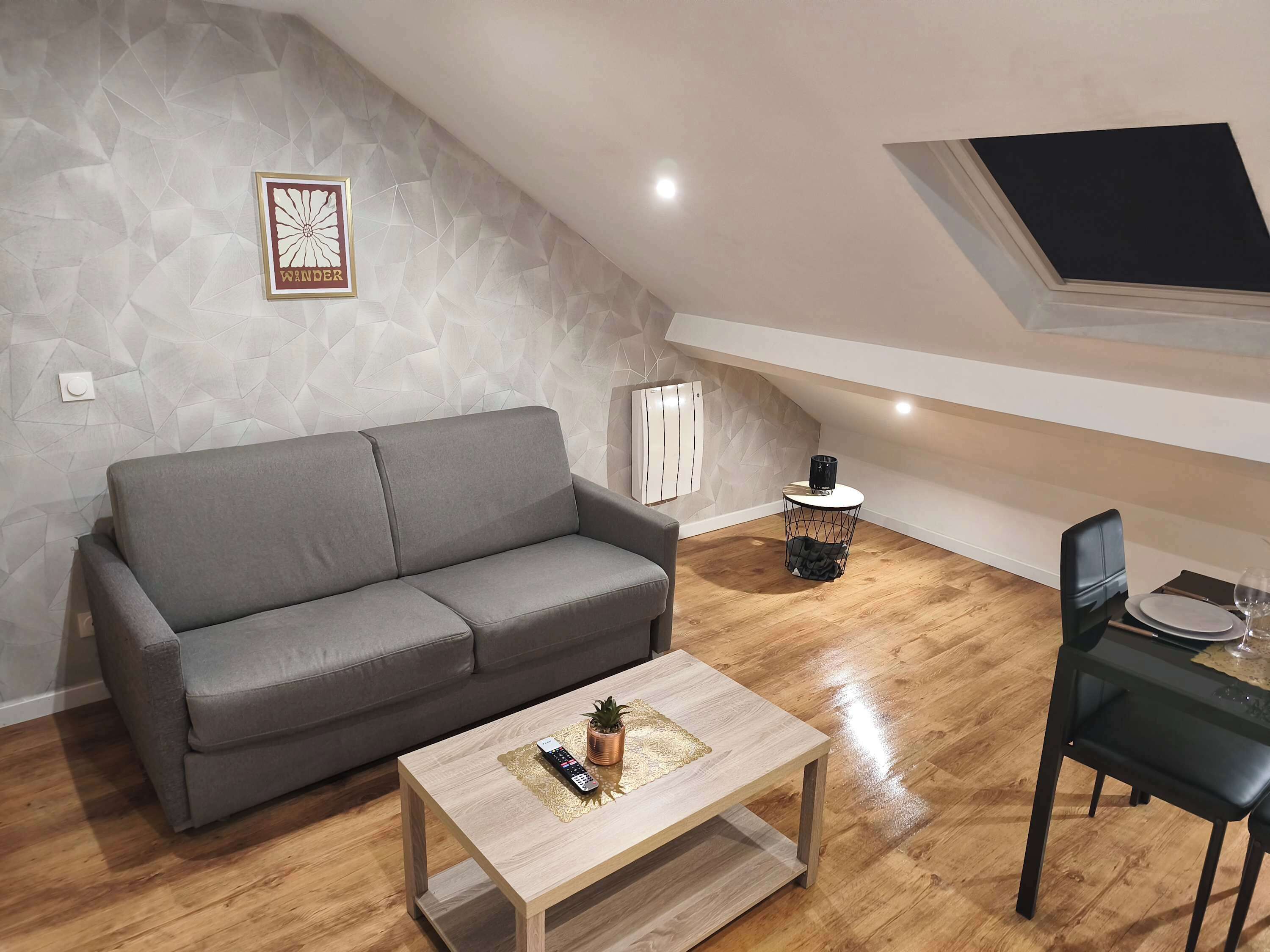 Un salon moderne avec un canapé gris, une table basse en bois sur laquelle se trouvent une télécommande et une petite plante, un fauteuil noir et une table à manger dressée avec une assiette et un verre.