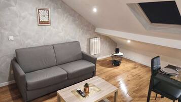 Un salon moderne avec un canapé gris, une table basse en bois sur laquelle se trouvent une télécommande et une petite plante, un fauteuil noir et une table à manger dressée avec une assiette et un verre.
