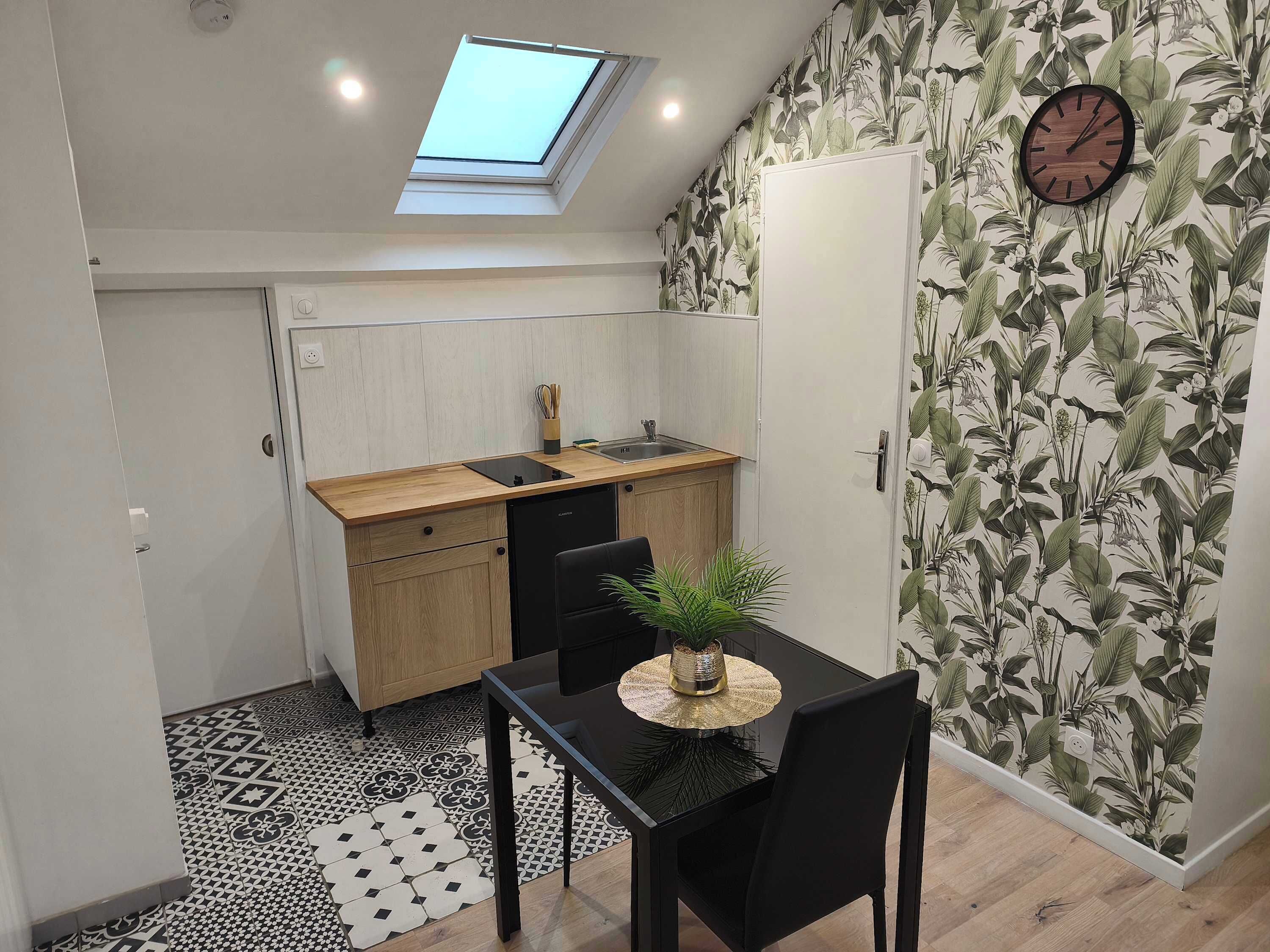 Une cuisine compacte dotée d’un puits de lumière, un coin repas avec une table et des chaises noires, et une horloge murale.