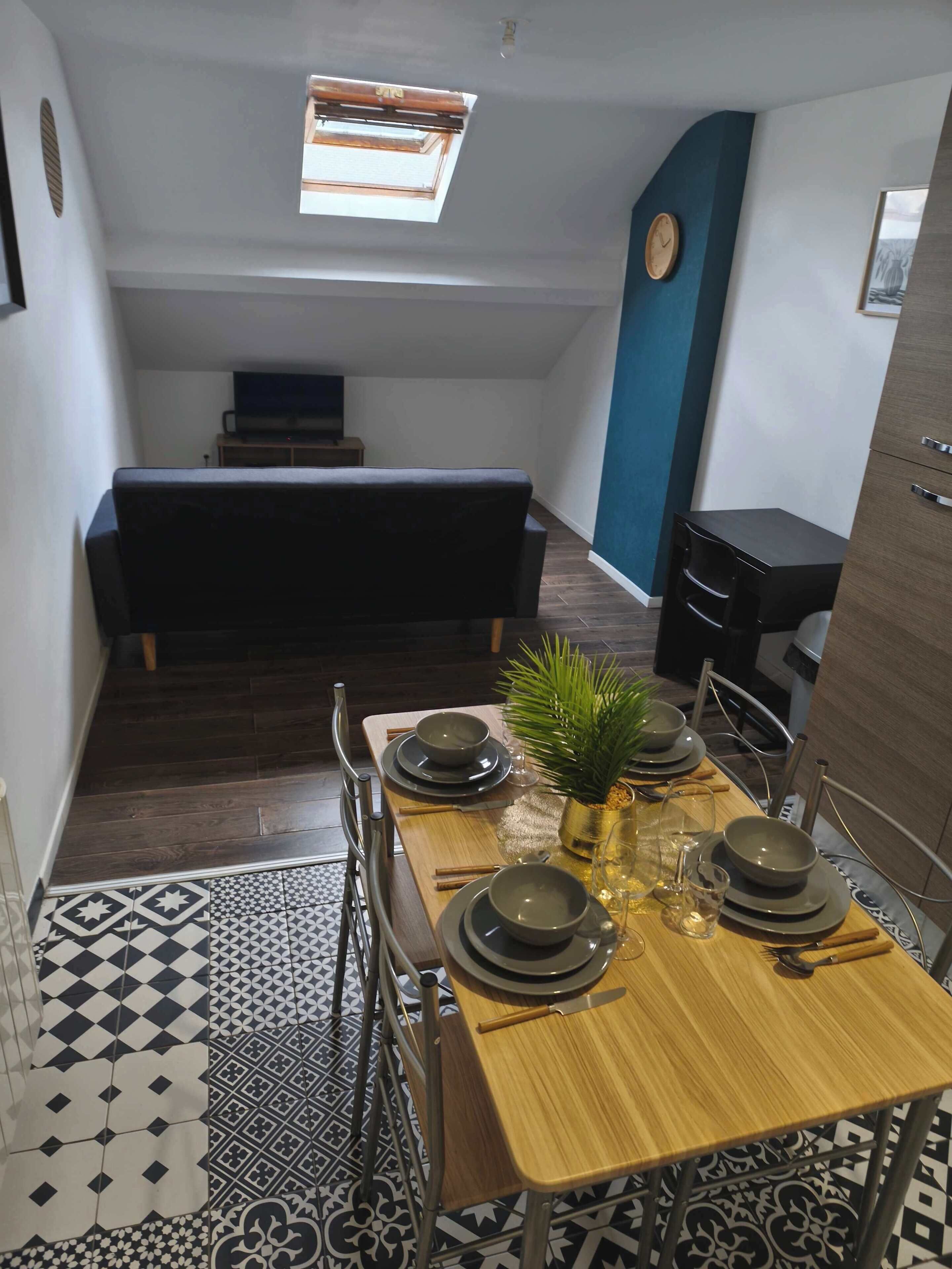 Un comedor con una mesa de madera preparada para cuatro personas, una pequeña planta en maceta y una pared azul con un reloj.