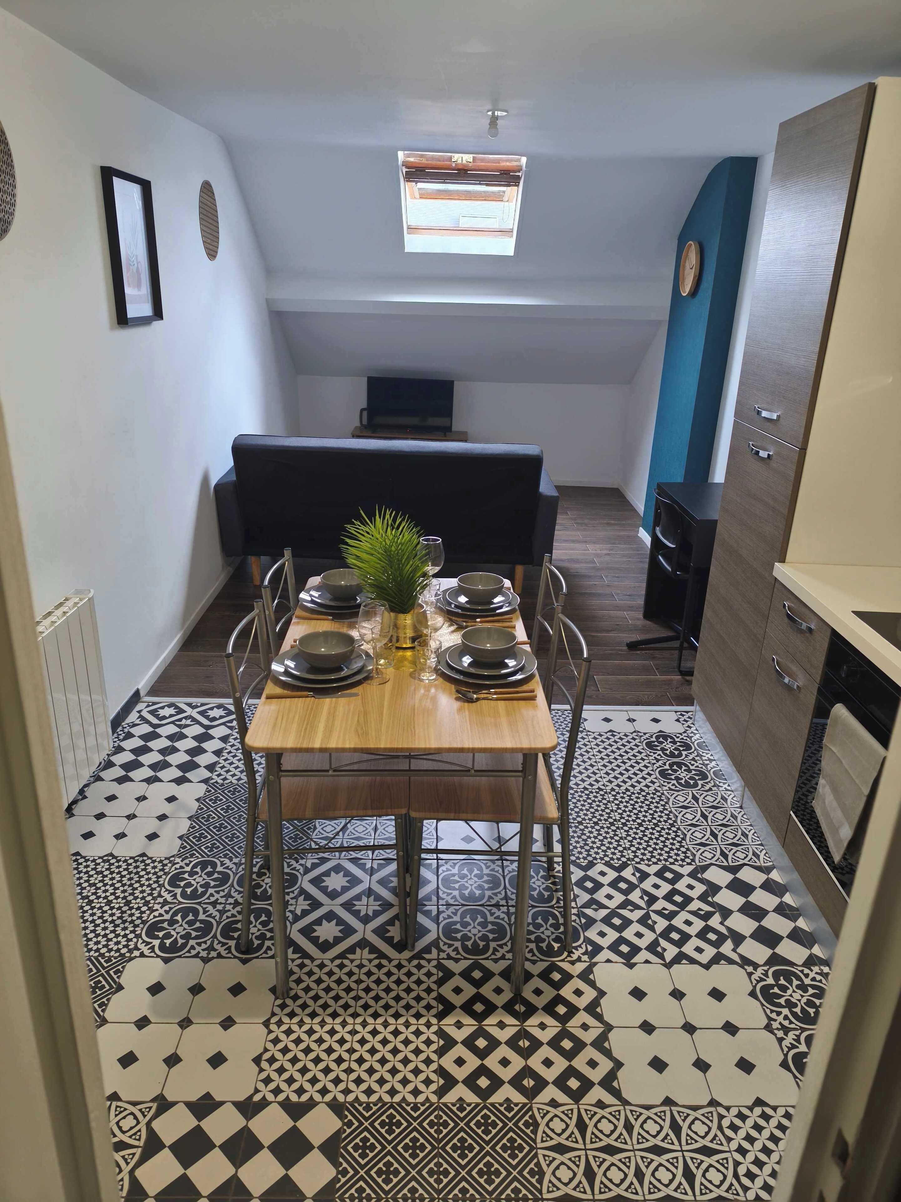 Una cocina compacta con comedor, que presenta un suelo de baldosas estampadas, una pequeña mesa de comedor con platos y una planta, y un televisor al fondo.