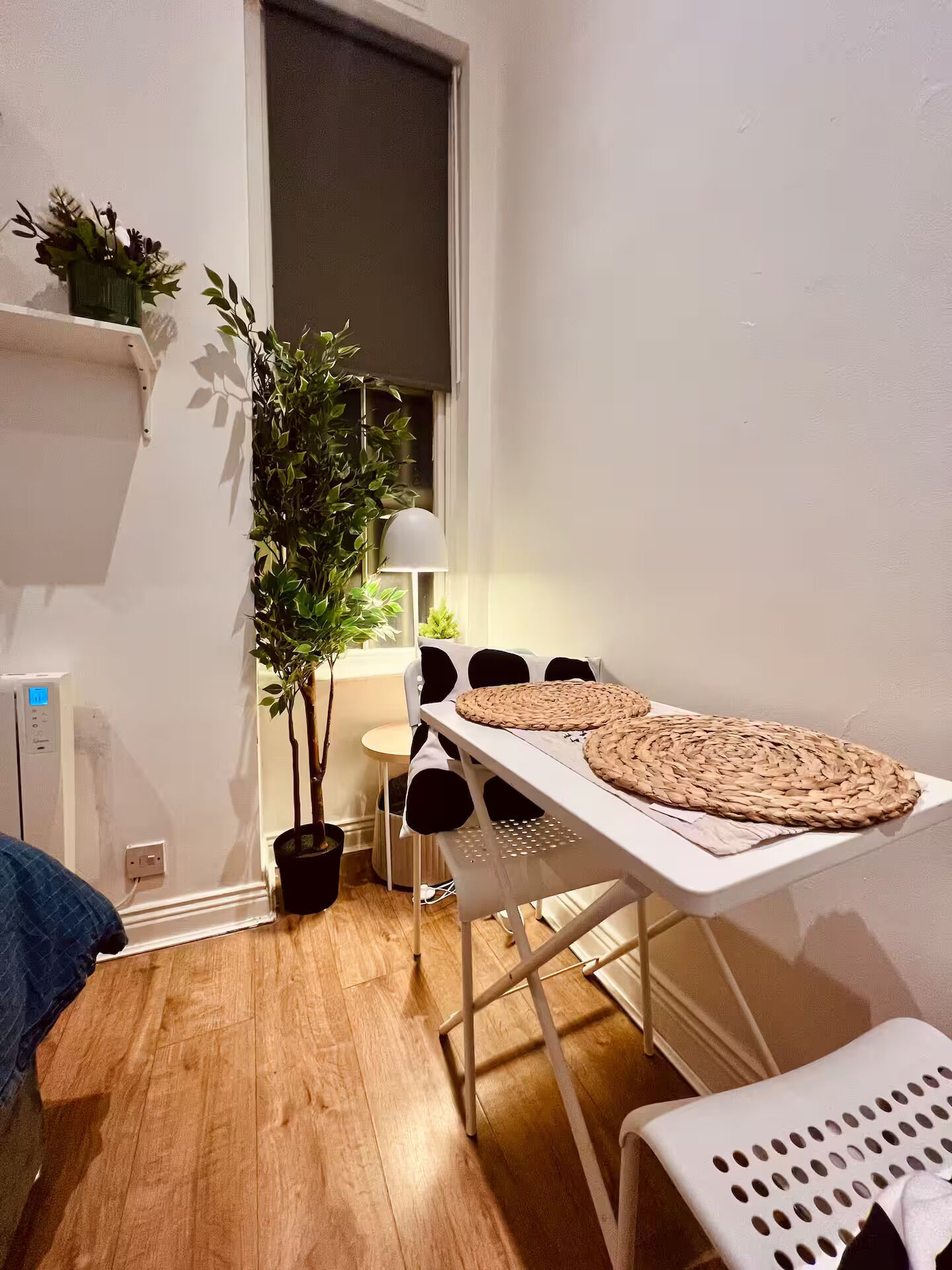 A small, well-lit room with a wooden floor, a white table with woven placemats, a potted plant, and a lamp.