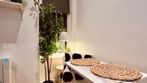 Une petite pièce bien éclairée, avec un plancher en bois, une table blanche surmontée de sets de table en fibres tressées, une plante en pot et une lampe.