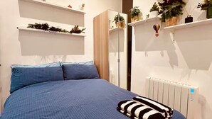 Une chambre avec un grand lit, des murs blancs et des étagères ornées de plantes et d’objets décoratifs.