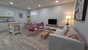 Una sala de estar moderna con un sofá gris, una mesa de centro y un televisor. Hay una zona de comedor con una mesa y sillas, y una cocina con armarios blancos.