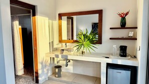 Une salle de bain moderne avec un lavabo blanc, un miroir, une plante en pot, une machine à café et un petit réfrigérateur.