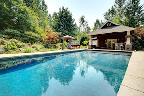 Une piscine aux eaux cristallines, dotée d’une terrasse en bois et d’un espace détente, le tout entouré d’une végétation luxuriante et d’arbres.