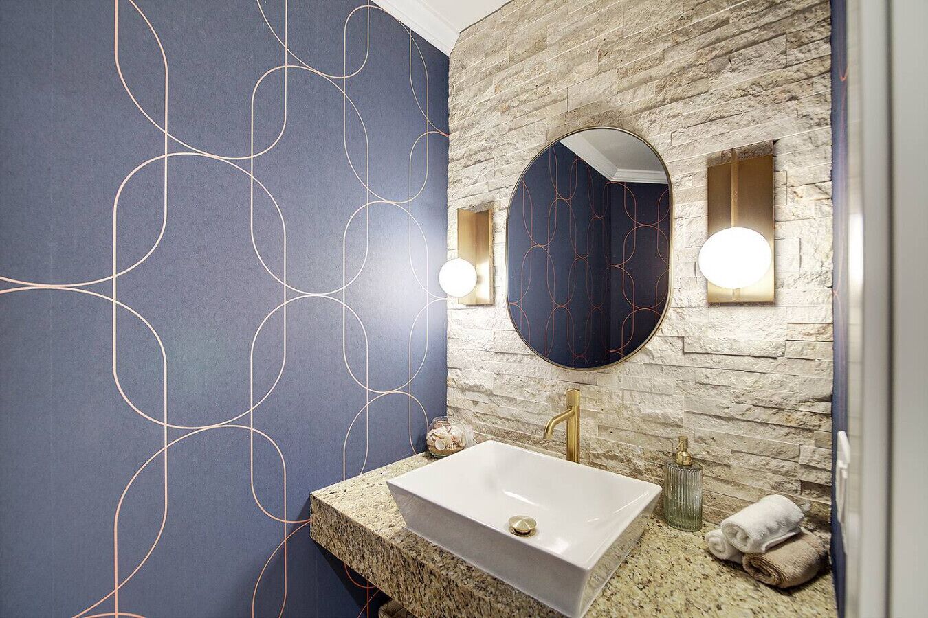 A bathroom with a stone wall, a blue patterned wall, a white sink, and a round mirror.