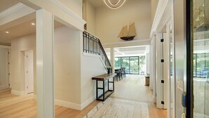 A spacious hallway with a wooden floor, a marble-patterned rug, a staircase with a ship model, and a view of a dining area with outdoor seating. - Luxury Wooded Estate With Pool (Sammamish)