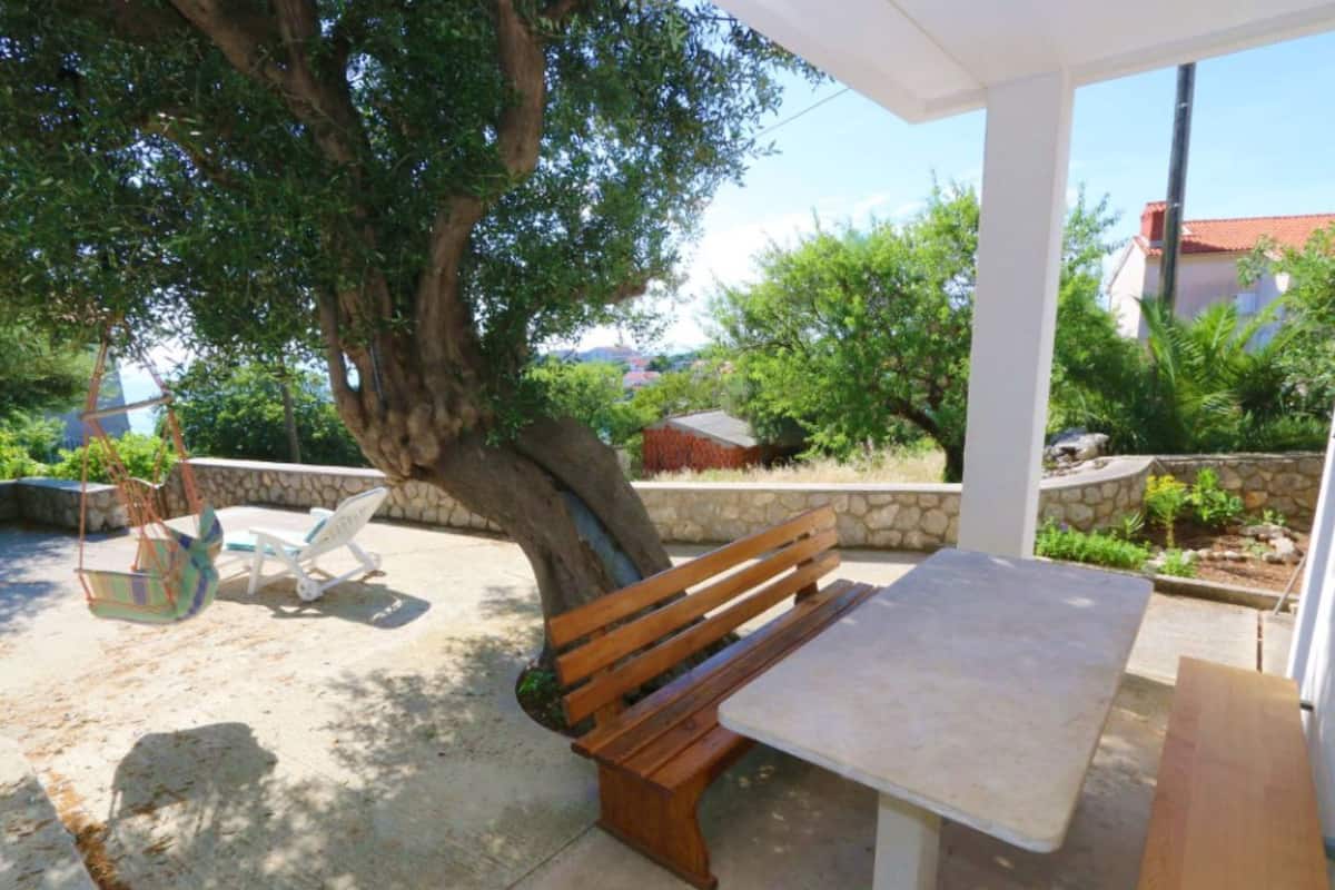A covered outdoor seating area with a wooden bench and a table, surrounded by greenery and a stone wall.
