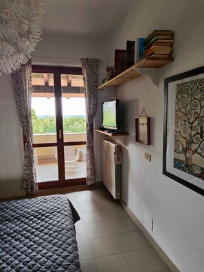 A bedroom with a bed, a television, a radiator, a window with curtains, and a wall-mounted shelf with books. - Farmhouse Tenuta Blueberry - Glicine near Milan-Malpensa (Cascinetta)