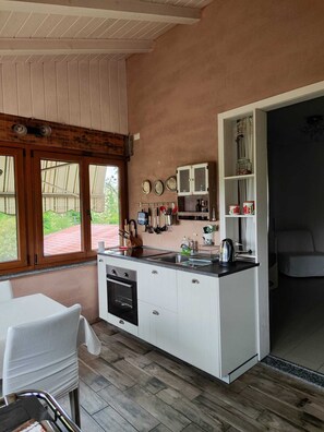 A compact kitchen with a built-in oven, a dining area with a white table and chair, and a large window offering a view of the outdoors. - Farmhouse Tenuta Blueberry - Glicine near Milan-Malpensa (Cascinetta)