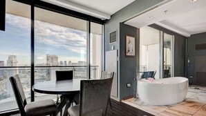 Una habitación de hotel moderna con un ventanal amplio que ofrece vistas a la ciudad, una mesa de comedor redonda y una bañera independiente.