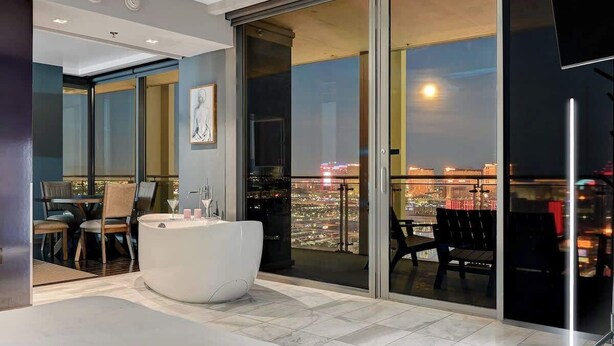 Un baño moderno con una bañera grande, un balcón con cerramiento de vidrio y vistas a la ciudad, y un comedor con sillas y una mesa.