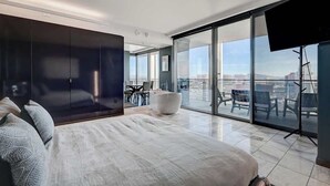 Un dormitorio moderno con una cama grande, una zona de estar con un reposapiés blanco y un balcón con vista a la ciudad.