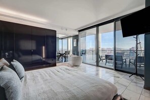 Un dormitorio moderno con una cama grande, una zona de estar con un reposapiés blanco y un balcón con vista a la ciudad.