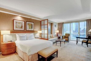 A hotel room with a large bed, a sofa, a small table, and a chair. There are two framed pictures on the wall, a window with curtains, and a view of buildings outside. - Signature Strip View Suite (Las Vegas)