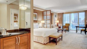 Une chambre d’hôtel avec un grand lit, un bureau, une chaise, un miroir et une vue sur la ville grâce à de grandes fenêtres.
