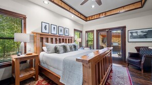 Une chambre avec un lit en bois, une table de chevet avec une lampe, une grande fenêtre avec des stores et une vue sur l’extérieur.