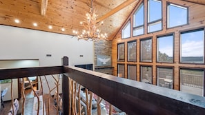 A spacious room with a high ceiling, wooden beams, and large windows offering a view of the outdoors. - New! Unbelievable Views! Gourmet Kitchen! Firepit! (Broken Bow)