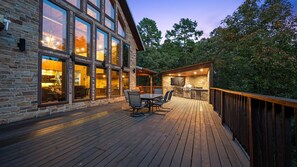 Une terrasse en bois avec des meubles d’extérieur, un bâtiment en pierre doté de grandes fenêtres et un espace barbecue.