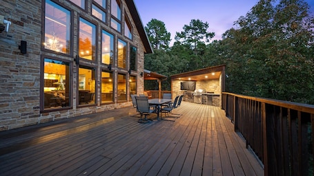 A wooden deck with outdoor furniture, a stone building with large windows, and a barbecue area.