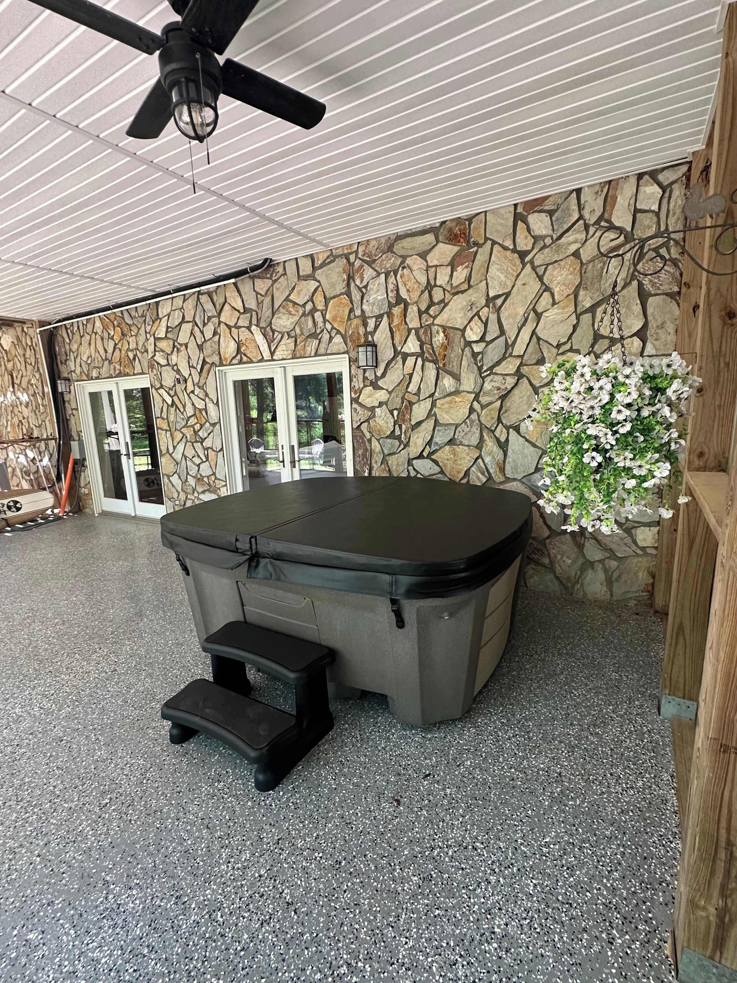 A covered outdoor area with a stone wall, a hot tub, and a ceiling fan.