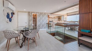 A modern kitchen with a glass island, a dining area with wireframe chairs, and a wooden accent wall. - Portamaris #301 - Luxury Penthouse by Carpediem (Aquiraz)