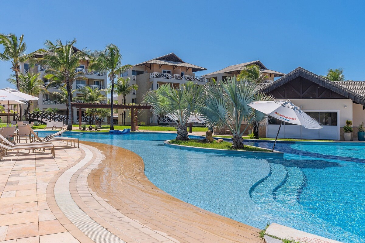 Ein Poolbereich einer Ferienanlage mit Palmen, Liegestühlen und einem Gebäude mit Balkonen.