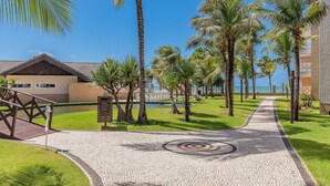 A paved pathway leading to a building with a tiled roof, surrounded by palm trees and a grassy area. - Portamaris #301 - Luxury Penthouse by Carpediem (Aquiraz)