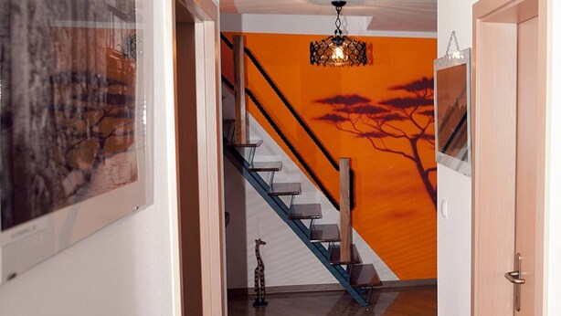 Una escalera con barandilla de madera y escalones de metal que conduce a una habitación con pared naranja y un mural de un árbol.