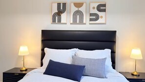 Een net opgemaakt bed met witte lakens en twee kussens, met aan weerszijden nachtkastjes met lampen, tegen een muur met abstracte kunst.