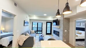 Ein modernes Wohnzimmer mit einer Couch, einem Couchtisch und einem an der Wand befestigten Fernseher. Es gibt zwei Fenster mit schwarzen Rahmen und einen Kamin.