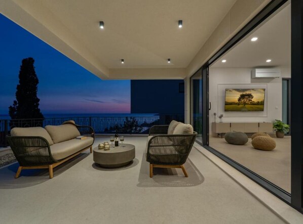 A modern outdoor living area with a sofa and chairs, a round table, and a view of the sea at dusk.