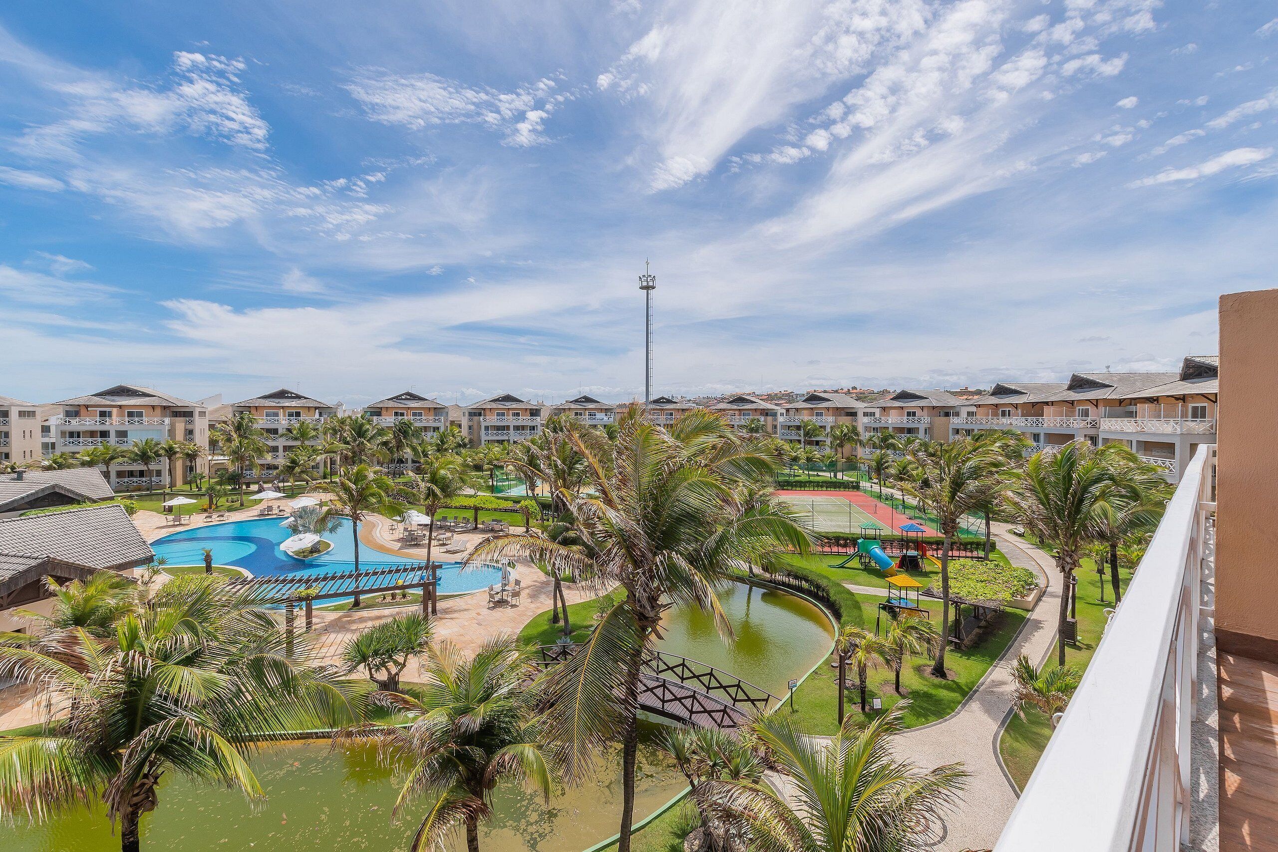 Un resort con varias edificaciones, una piscina, palmeras y cielo despejado.