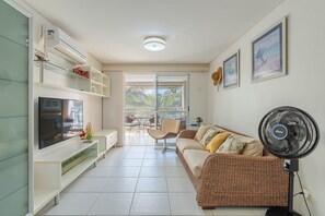 A modern living room with a flat-screen TV, a wicker sofa, a standing fan, and a glass door leading to an outdoor area.