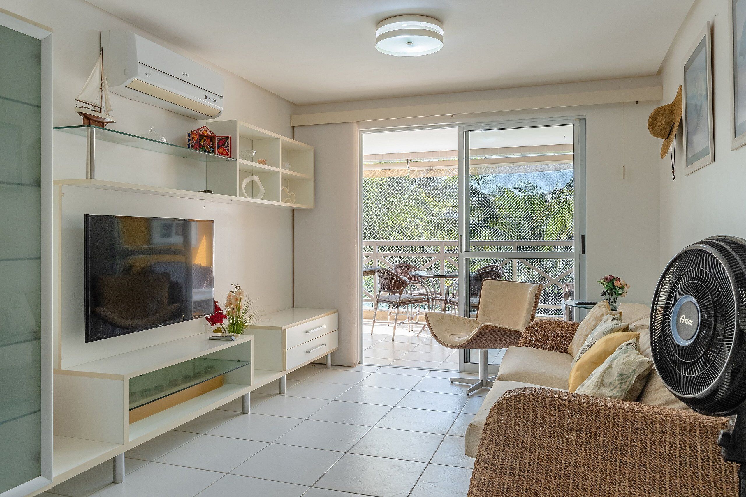 A modern living room with a flat-screen TV, white shelving, wicker furniture, and a large fan.
