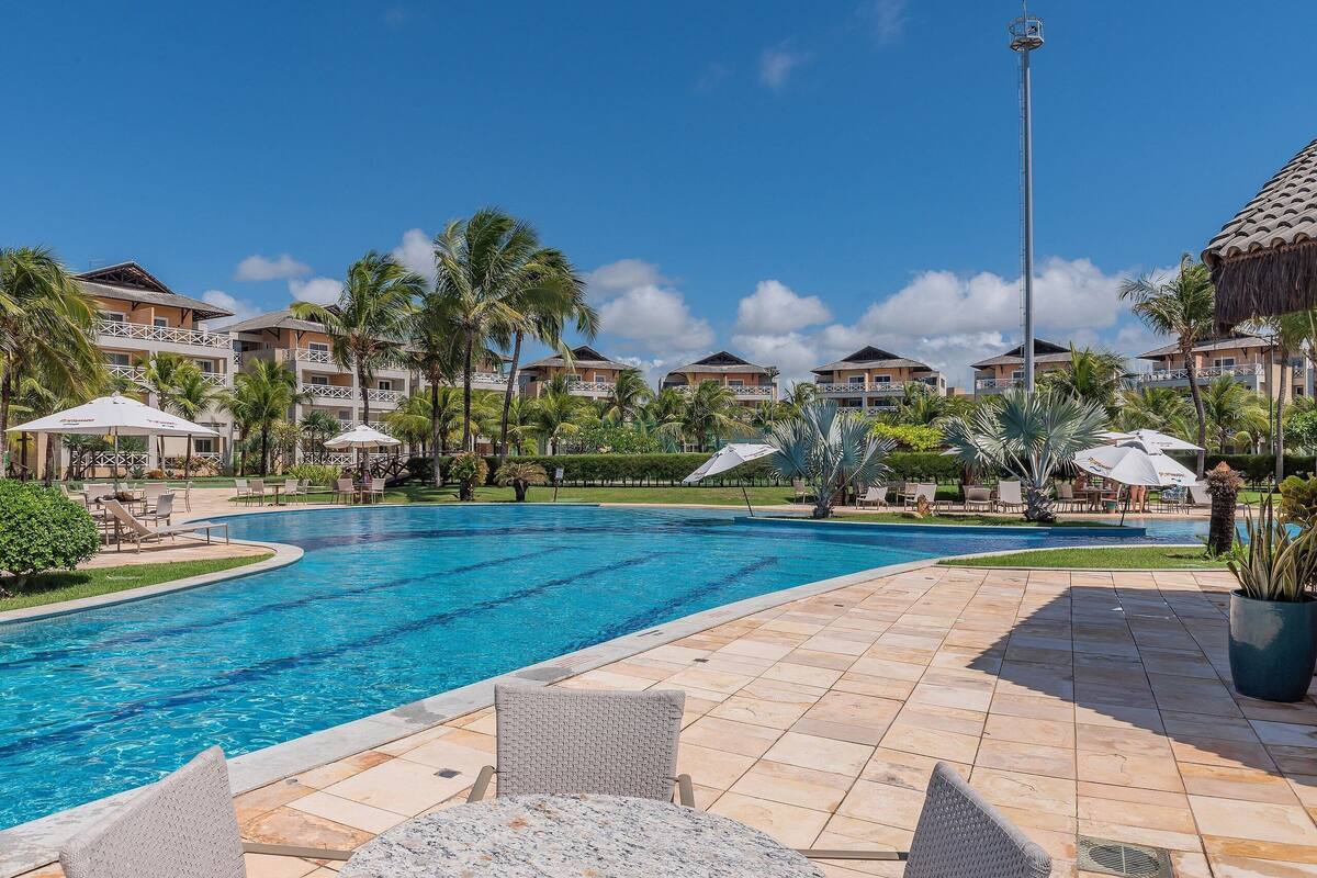 A swimming pool with lounge chairs and umbrellas, surrounded by hotel buildings and palm trees.